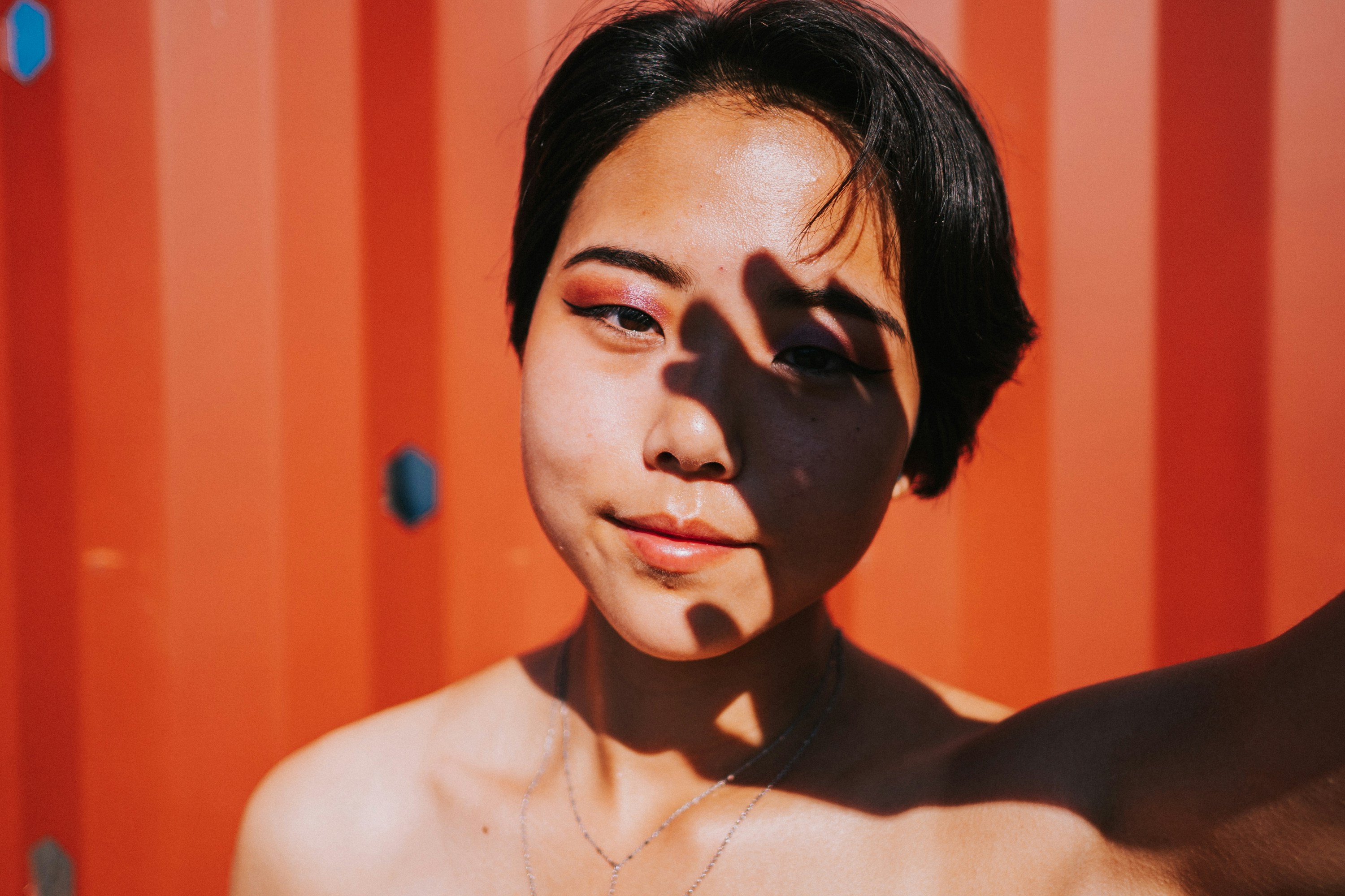 An image of a young woman with short black hair standing in front of a red garage door. The woman's face is in shadow, and...