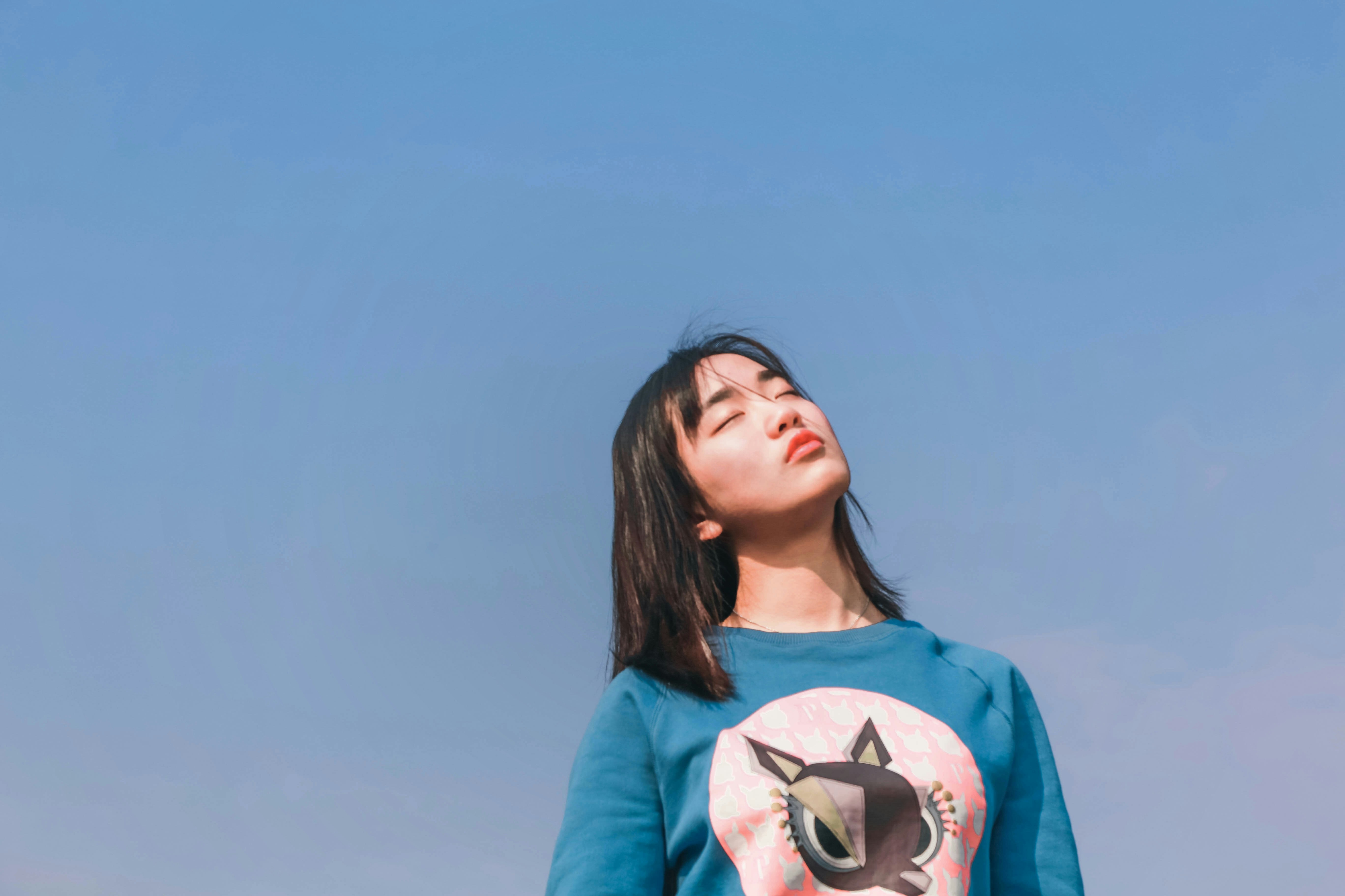 An image of a woman with her eyes closed and head tilted back, enjoying the sun, wearing a teal-blue sweater with a whimsi...