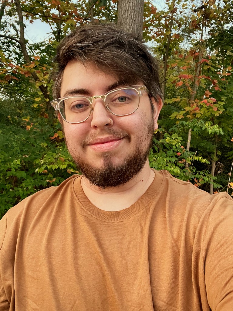 Sam, a mixed race transgender man with olive toned skin, smiles at the camera with fall foliage behind him.